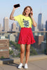 Woman in a yellow shirt and red polka dot skirt, holding a phone and wearing headphones, with a city skyline background.
