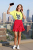 Woman in a yellow shirt and red polka dot skirt, holding a phone and wearing headphones, with a city skyline background.