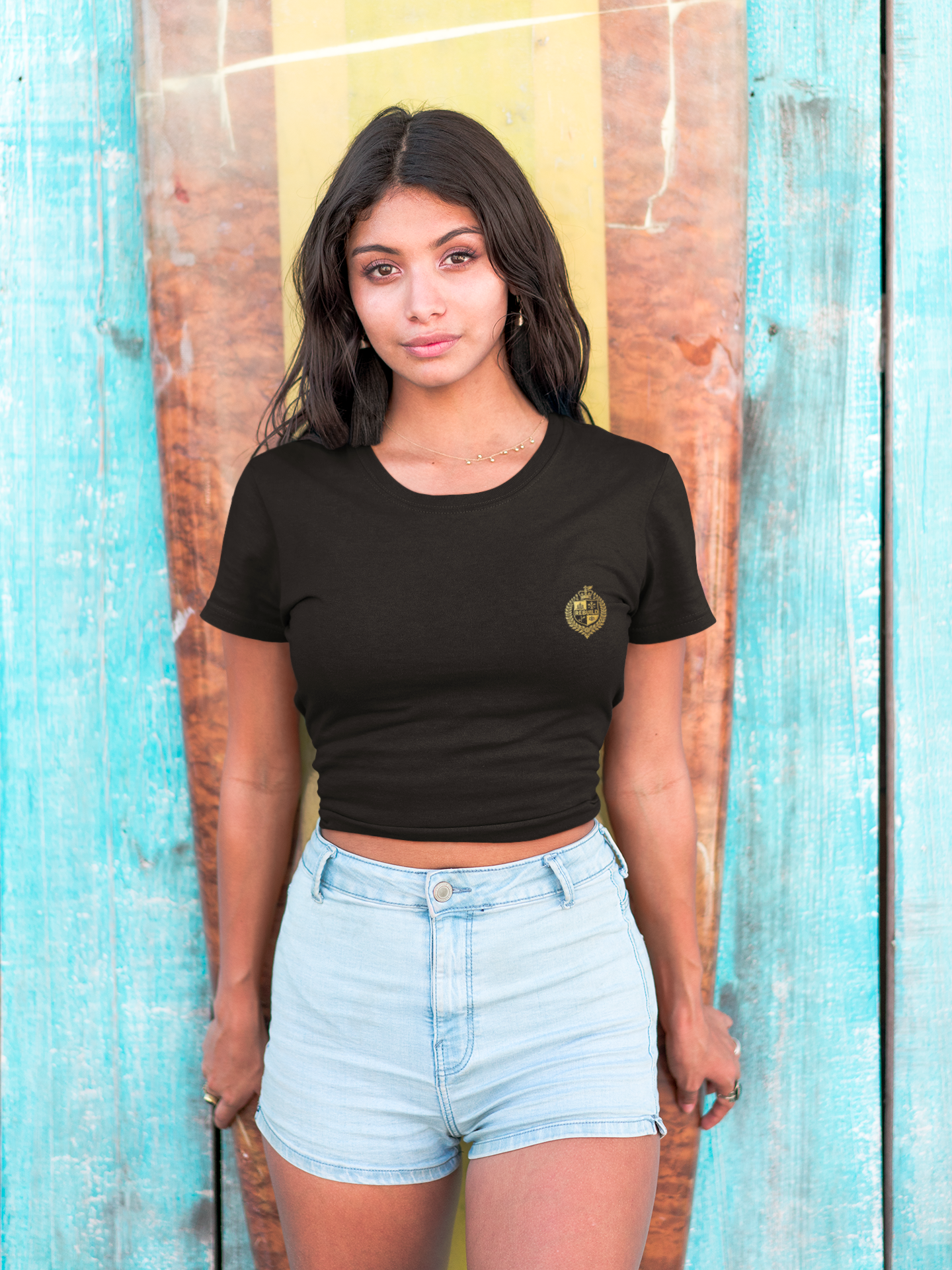Woman in black crop top and light denim shorts standing by surfboard against a blue wooden wall