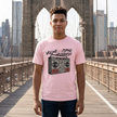 A man wearing a light pink graphic t-shirt stands on a wooden bridge with a city skyline and suspension bridge structure in the background. The t-shirt features a black and red boombox graphic with the text 'VOLUME OF COPING MECHANISM'.
