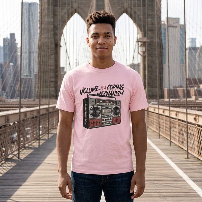 A man wearing a light pink graphic t-shirt stands on a wooden bridge with a city skyline and suspension bridge structure in the background. The t-shirt features a black and red boombox graphic with the text 'VOLUME OF COPING MECHANISM'.