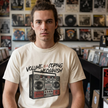 A man with long hair stands in a record store wearing a cream-colored graphic t-shirt. The t-shirt features a black graphic of a boombox with the text 'VOLUME IS A COPING MECHANISM' printed above it.